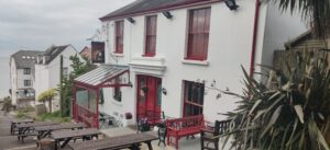 The outside of the Clarendon pub. There are benches outside and a little porch area with a little roof. In the distance you can see the sea.