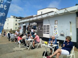 The outside of the Boat House in Sandgate. There are tables and chairs with people sitting outside on a sunny day. The tables have daffodils on them and people are chatting.